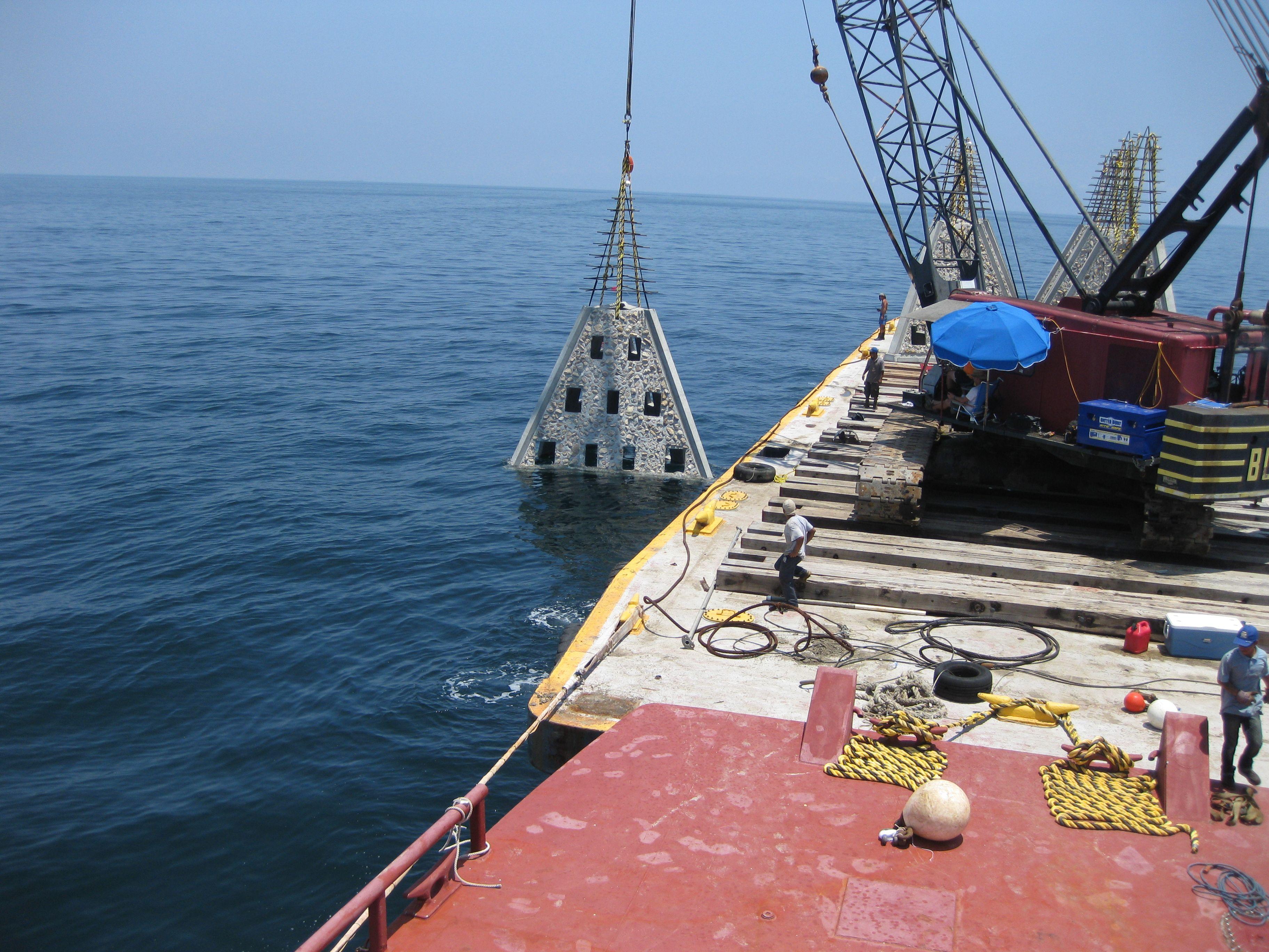 Crane lowering a pyramid-shaped reef module from a barge